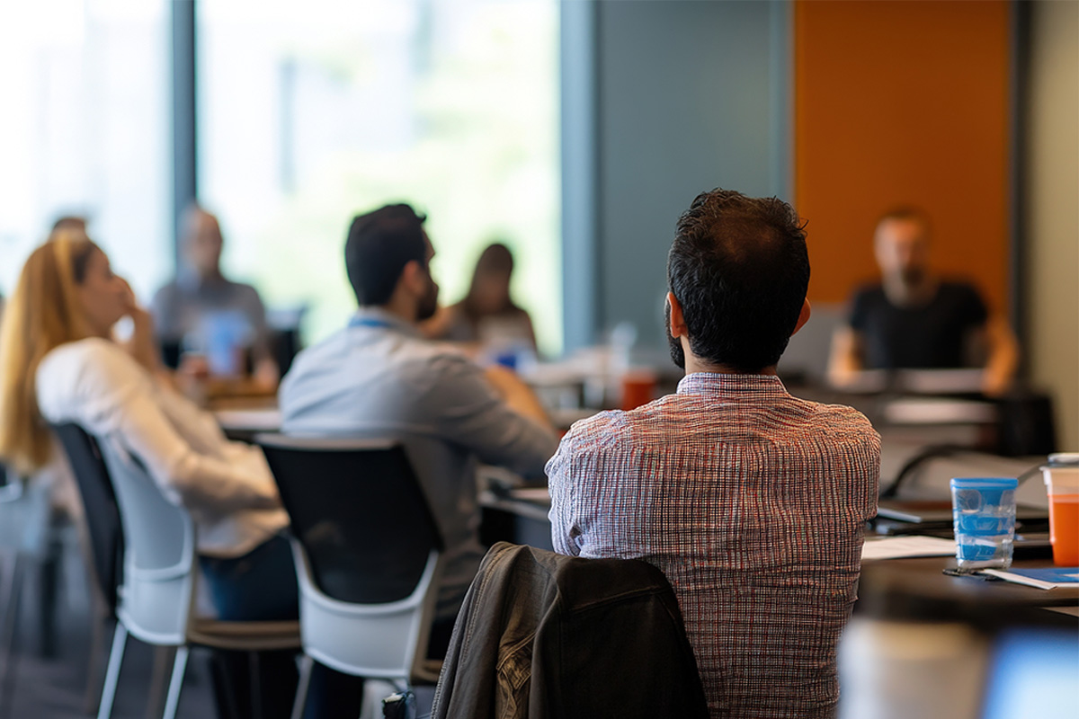 A group of adults seated around a conference table in a meeting or workshop, listening to a speaker at the front of the room.