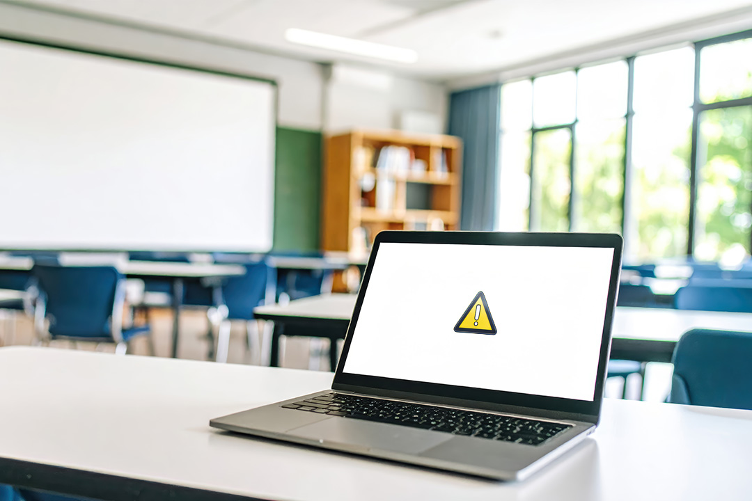 laptop with a blank screen sits on a desk in a classroom, displays a yellow warning screen with an exclamation mark inside a triangle.