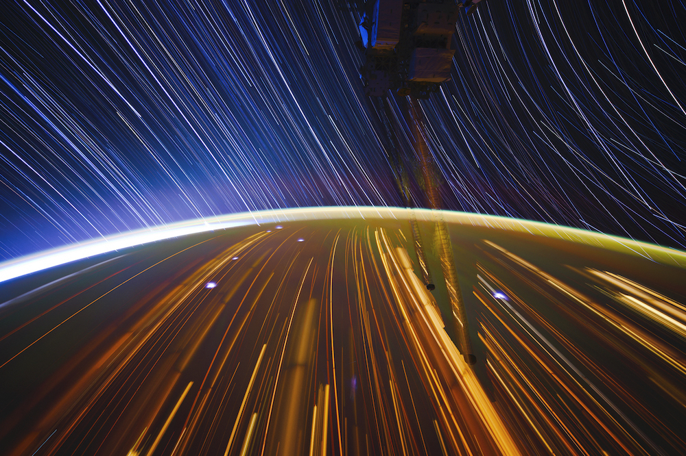 Photographic image of earth taken from the International Space Station with star trails of color and an aura of light along the curvature.