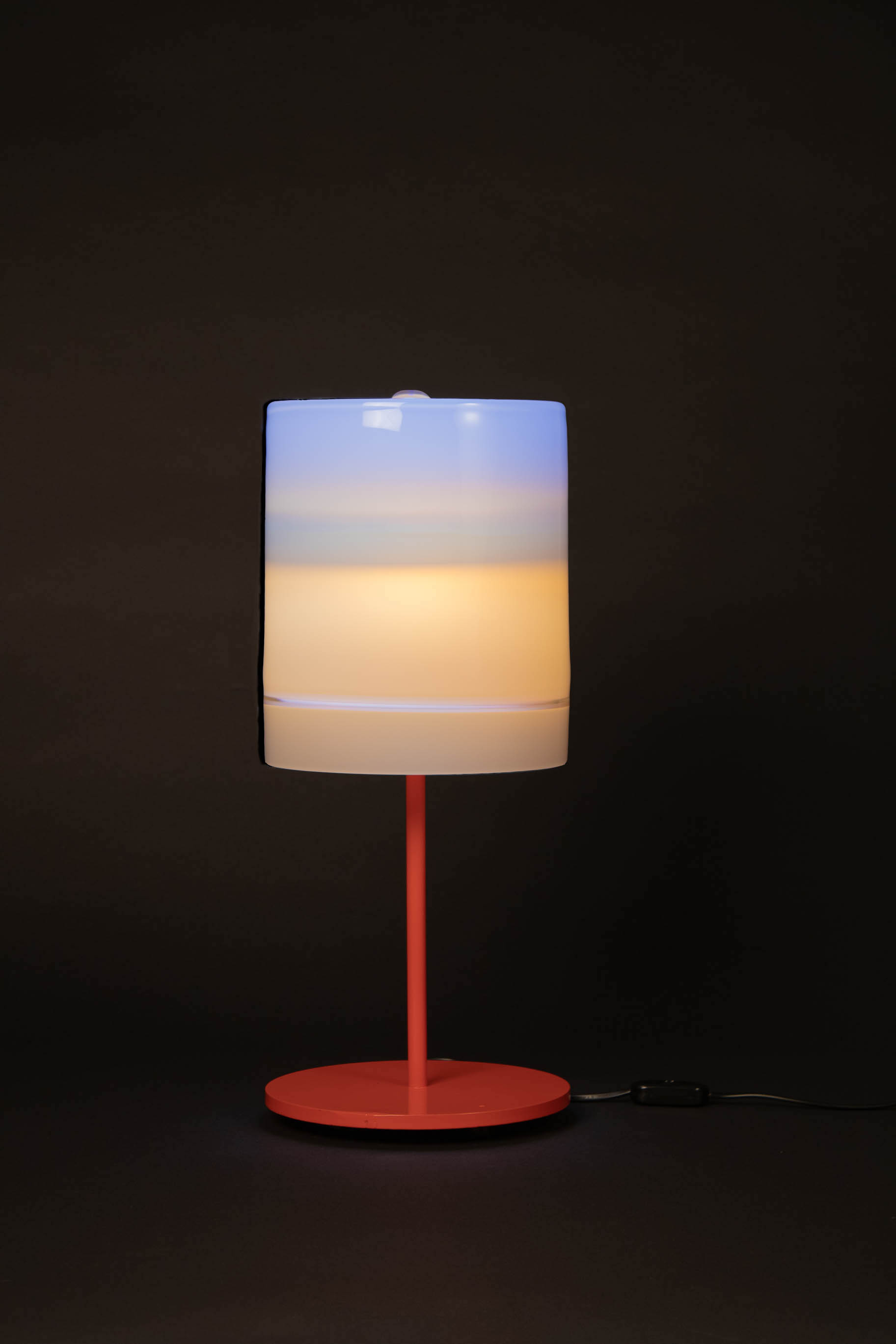 a lamp with a red metal base and white cylindrical shade against a black background.
