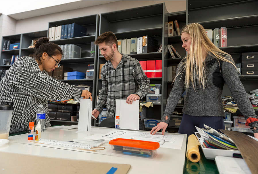 a color photograph of three college students working together on a studio work table with paper, rulers, and paper.