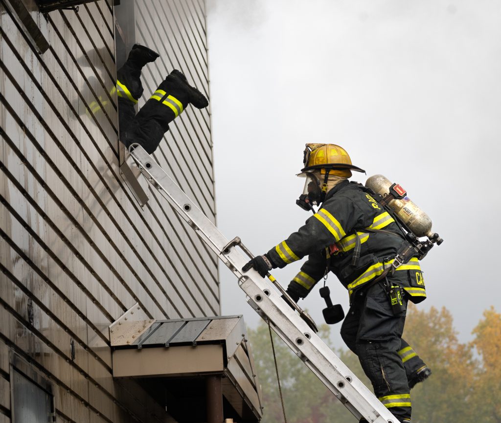 a color photo of a firefighter climbing an angled ladder toward an upper story window with smoke pouring out.