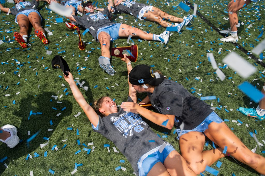 a color photograph of a group of lacross players lying on the ground in celebratory posed with a blizzard of blue confetti overhead.