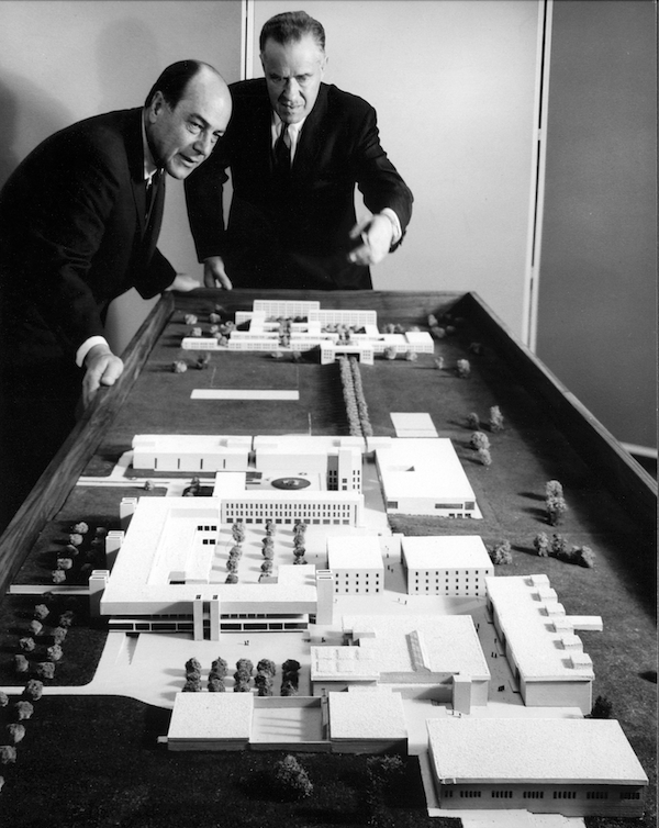 a black and white photograph of two men in suits and ties looking over a large scale model of a college campus.