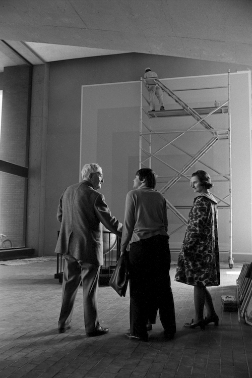 a black and white photograph of a group of three people standing in front of a large scale wall mural having a conversation. 