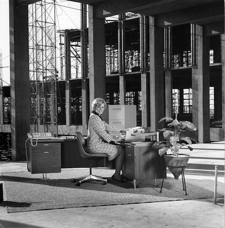 a black and white photograph of a woman in a professional work suit seated at an office desk in the midst of a full building construction site.