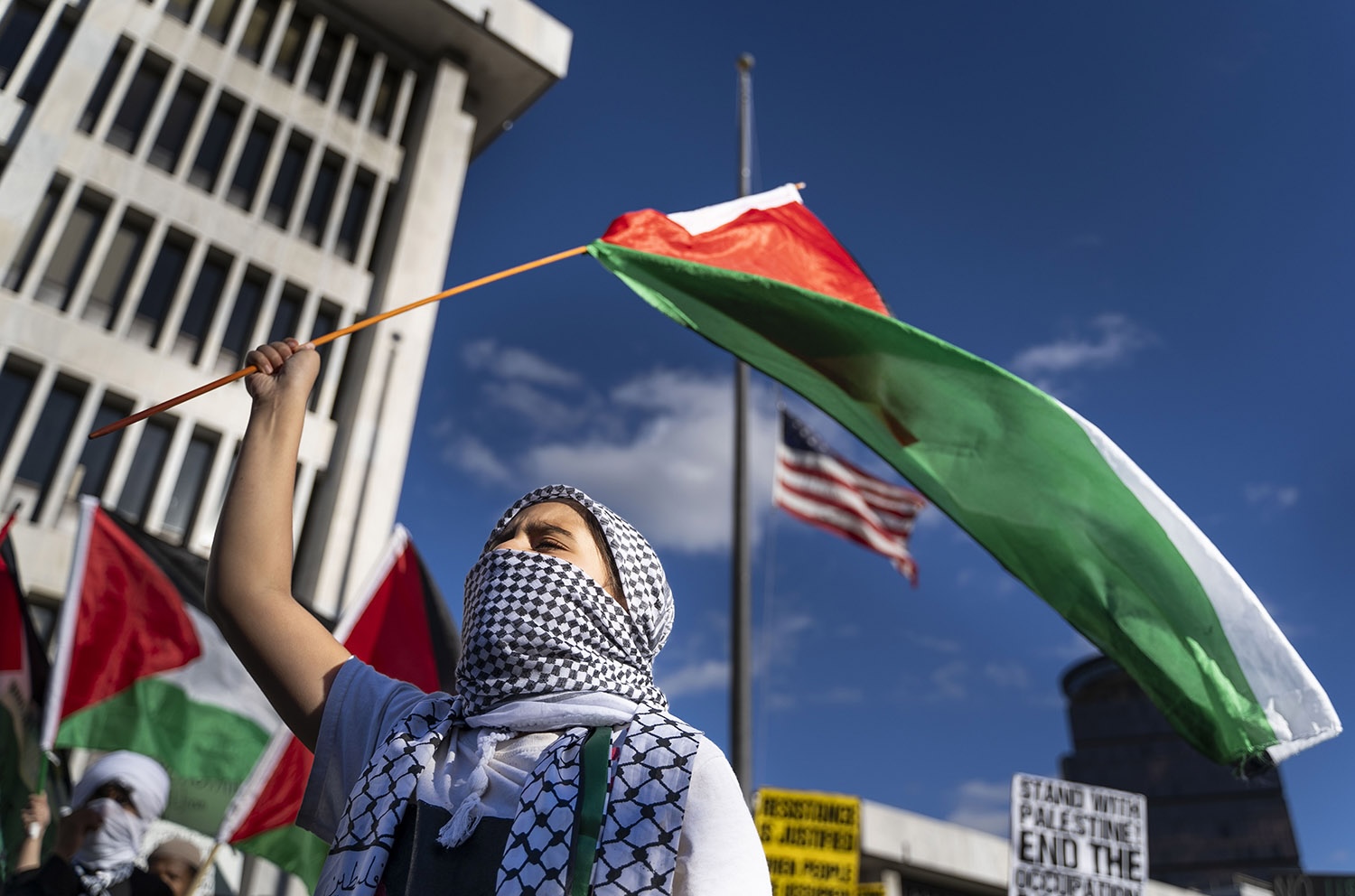a color photograph of a protestor with their head wrapped and face covered in checkered cloth holding a Palestinian flag on a stick overhead with the US flag in the background.