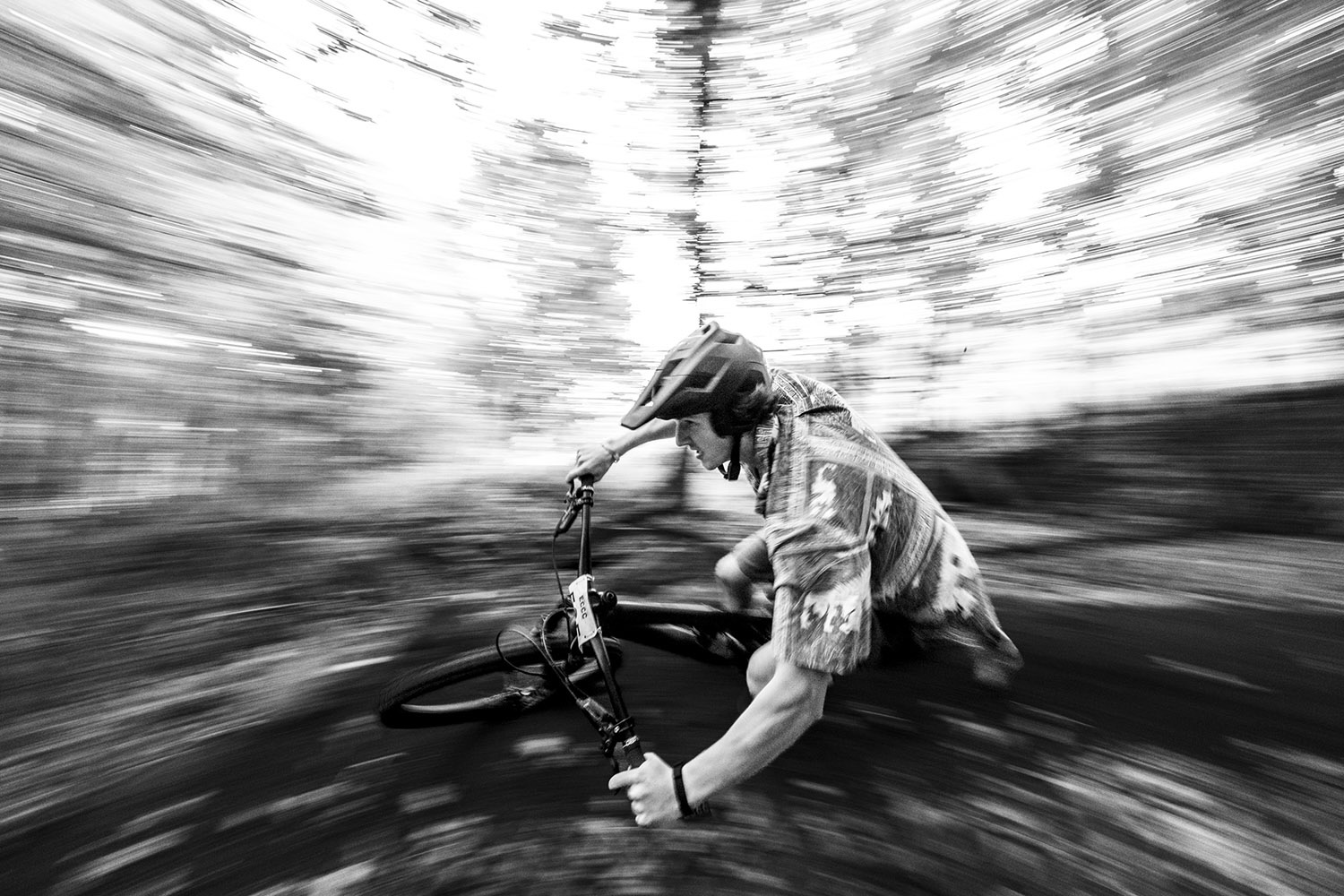 a black and white photograph of a racing bike rider in full motion heading around a banked surface, gripping the handle bars and the background in a motion blur.