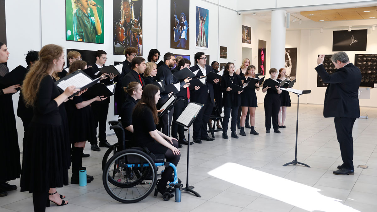 A choir of diverse singers, including a person using a wheelchair, performs in a gallery while a conductor leads them.