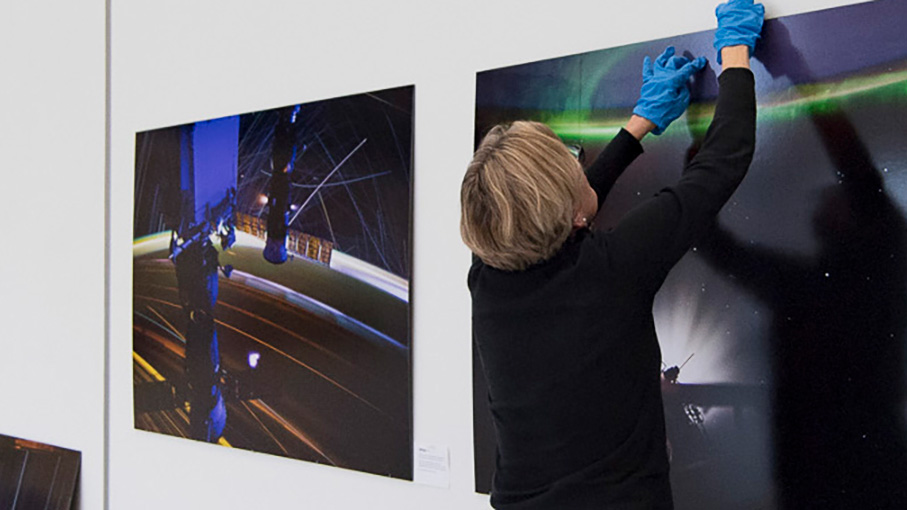A person wearing blue gloves installs a large photograph of an aurora and telescope equipment on a white gallery wall.