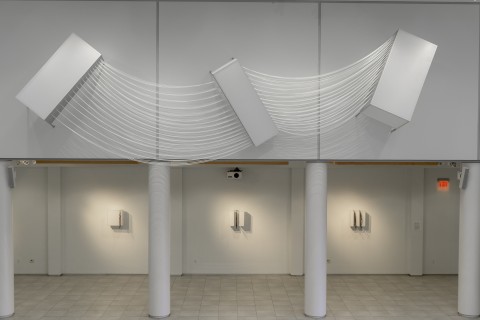 View of three white rectangular sculptural forms mounted on the upper wall with three smaller white box sculptures on the lower wall 