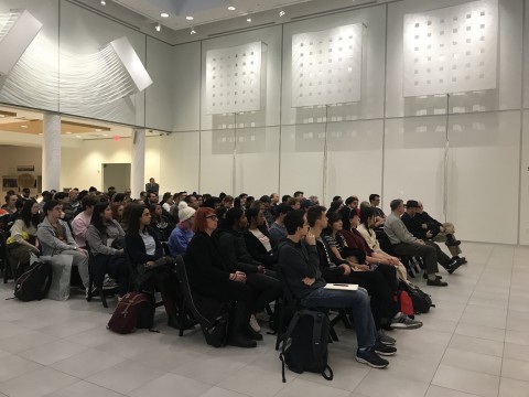 Image of audience seated in lecture style in gallery setting with white dimensional sculptures mounted on walls. 