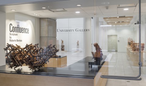View through the main glass window and door entrance to gallery with text on wall ' Confluence, Sculpture by Roberto Betoia'. Wood stacked form sculptures in foreground and background.