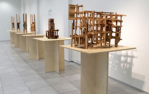 View of series of six stands each displaying a wood stacked form sculpture.