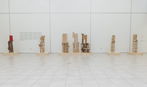 View across gallery floor to large white exhibition wall. Seven floor standing vertical sculptures displayed on low stands. 