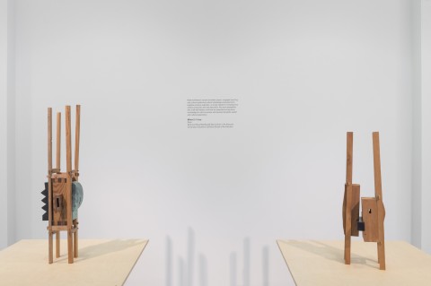 Two slender vertical sculptures with four long thin wood supports with wooden assemblage of wood blocks in the center. Exhibition text on wall.
