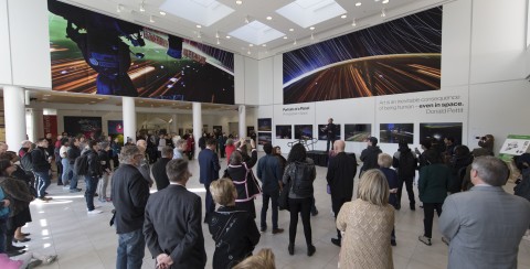 Full view of gallery with large gathering of people looking at 30 foot photo murals mounted on the second level of the walls.
