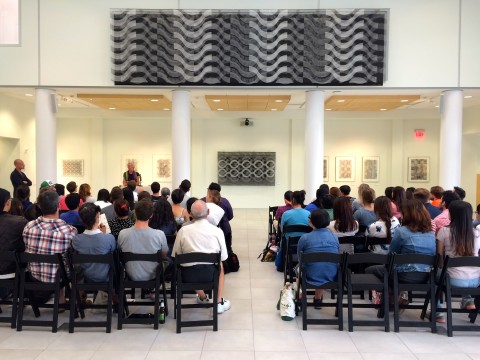 View of gallery installation - with audience of students seated in black chairs facing wall with large horizontal black and white wall hanging on upper level above lower arcade with undulating pattern.