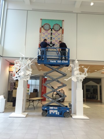 Image of two men standing in a hydraulic scissors lift while hanging one large rectangular painting on the upper wall.