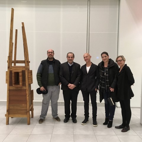 Group of five people standing next to Floor standing vertical sculpture with four long thin wood supports with wooden assemblage of wood blocks in the center. 