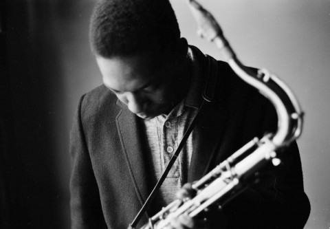 Black and white photo portrait of musician John Coltrane holding a saxophone and leaning, head bowed toward the camera.