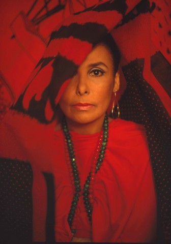 Color photo portrait of musician Lena Horne full on facing the camera with piercing dark color eye with a sash over the other eye, wearing a red dress, two strands of beads around her neck.