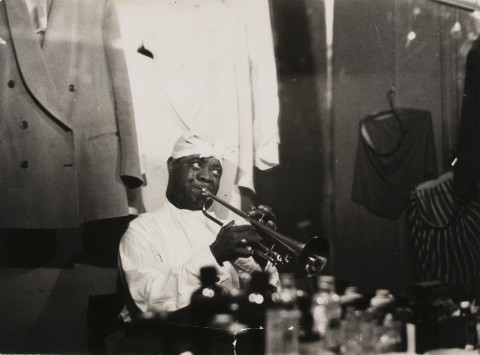 Black and white photo portrait of musician Louis Armstrong in his dressing room playing a trumpet with a white kerchief wrapped on his head tied with a central knot and two formal men's suits on hangers on either side. 