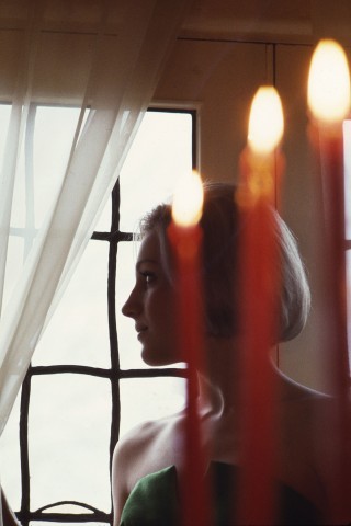 Color photo image of a young woman in profile with tall burning candles in foreground and panned window grid as a backdrop.