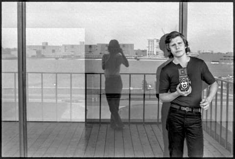Student holding a camera in a casual pose against a plate glass window with a reflection of the person taking the photo