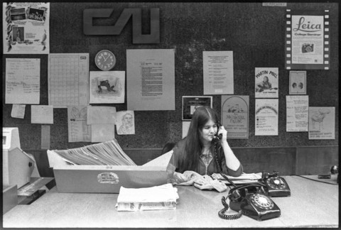 Image of a young woman answering a rotary phone at a reception desk in the Student Union