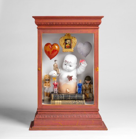 Alter like barn red wooden box with glass cover holding a white baby form with spikes protruding from forehead posed onto of 2 antique books and hearts dangling on either side