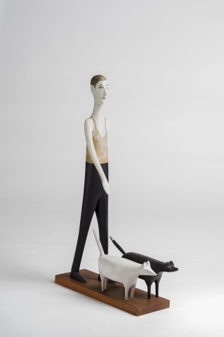 Standing carved wood sculpture of a tall and thin man in a walking stride holding a leash with one black dog and one white dog