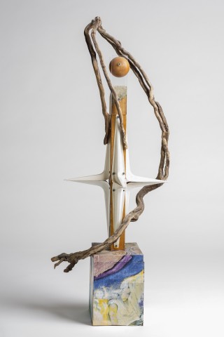 Standing sculptural form on a painted cube base - a central verticle core with metal protrusions and a found driftwood branch arching over the top with a single orange.