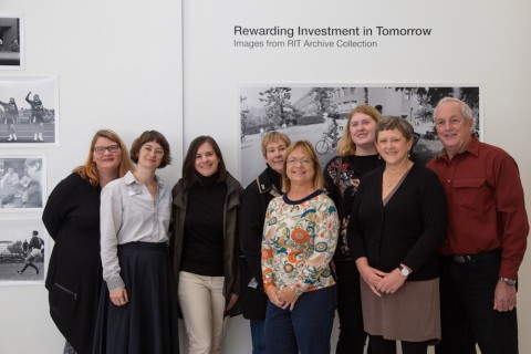 Group shot of eight people standing by the gallery exhibition wall text - Rewarding Investment in Tomorrow - 50 Years on the Henrietta Campus