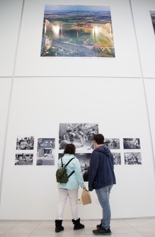 View of two story walls in gallery with two people looking at photos on the walls with one hung on the upper wall