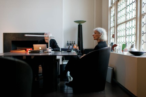 a view inside an artist studio with a wall of windows on the right and two people sitting at a black table.