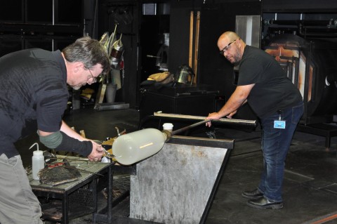 two people in a glass blowing studio working on a molten catch of white glass.