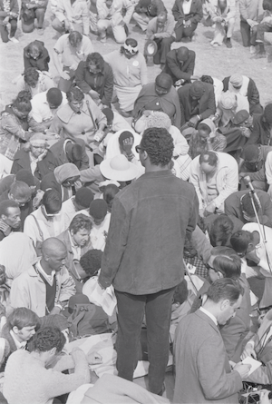 a black and white photograph of a large seated crowd of people with one person standing up facing the crowd.