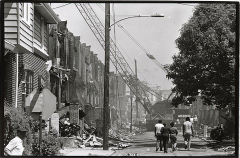 a black and white photograph of an urban street, a bombed out row of houses on the left and a group of four people walking away from the camera down the center of the street.