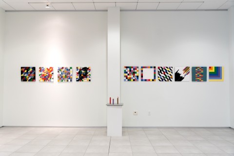 a display of colorful grid themed square paintings hung in a horizontal line on a vast white gallery wall.