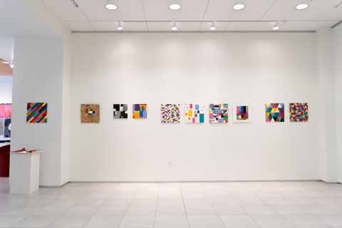 a display of colorful grid themed square paintings hung in a horizontal line on a vast white gallery wall.
