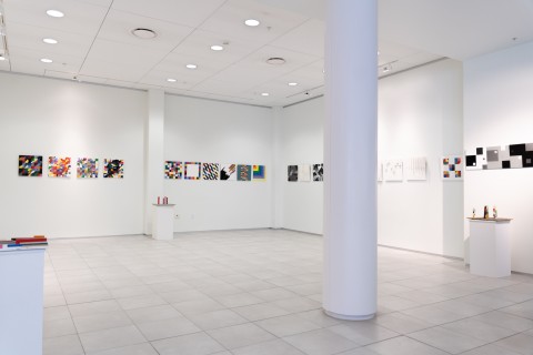 a display of colorful grid themed square paintings hung in a horizontal line on a vast white gallery wall.