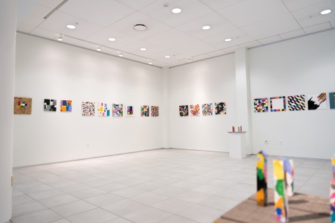 a display of colorful grid themed square paintings hung in a horizontal line on a vast white gallery wall.