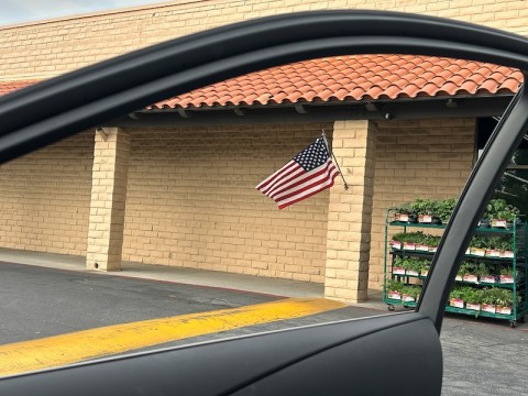 image taken from the interior of a car through the open passenger door of an American flag fluttering on a brick post. 
