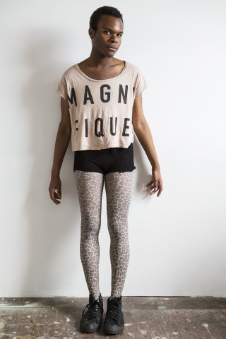 a color photograph of a person wearing a white t-shirt with bold graphic letters and black shorts leaning against a wall.