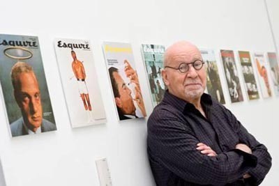 Photographs image of George Lois leaning onto a wall with copies of his designs mounted on the gallery walls 