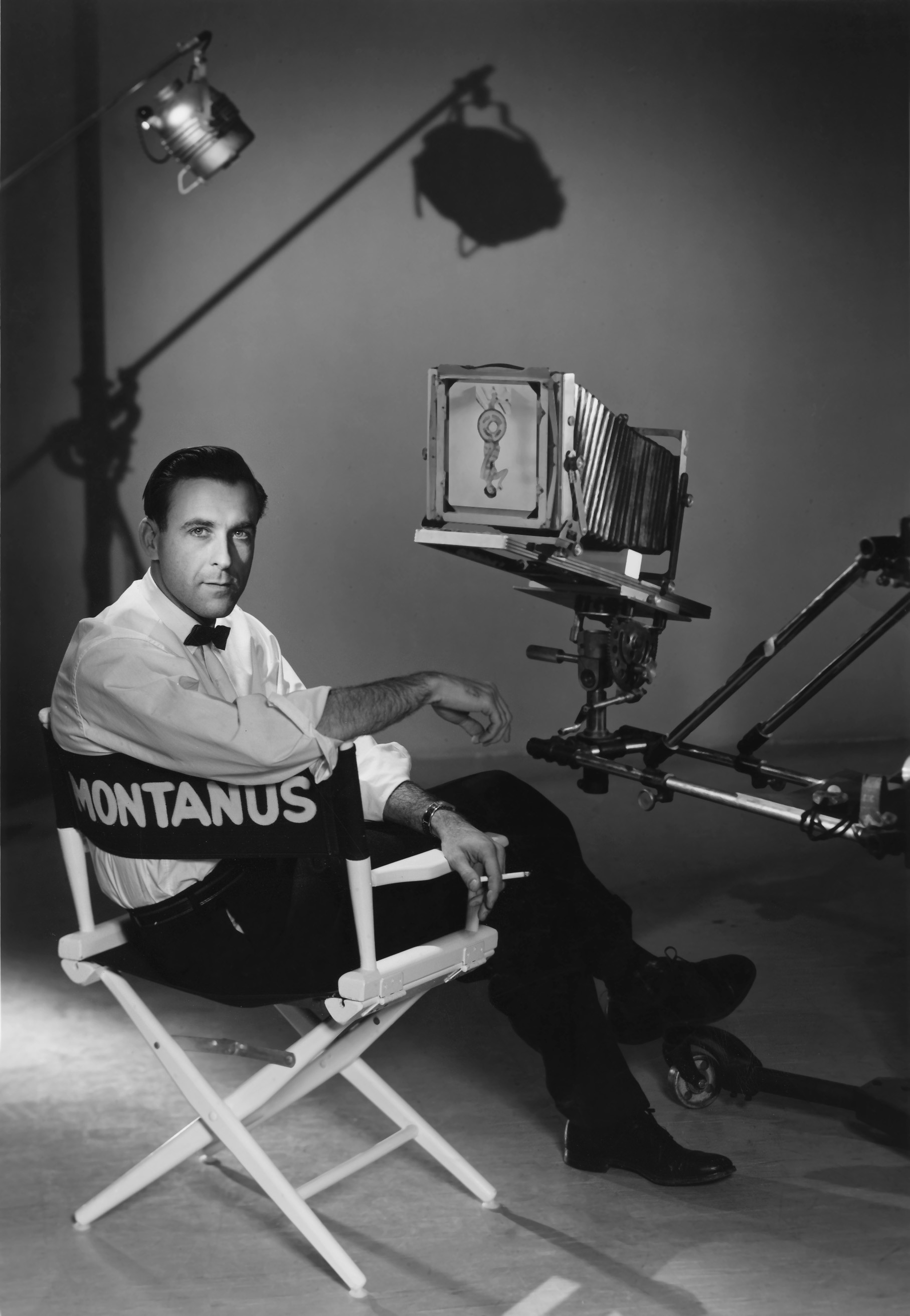 Photographic portrait of Neil Montanus wearing a white shirt and bow tie while seated in a director's chair with photographic equipment around him.