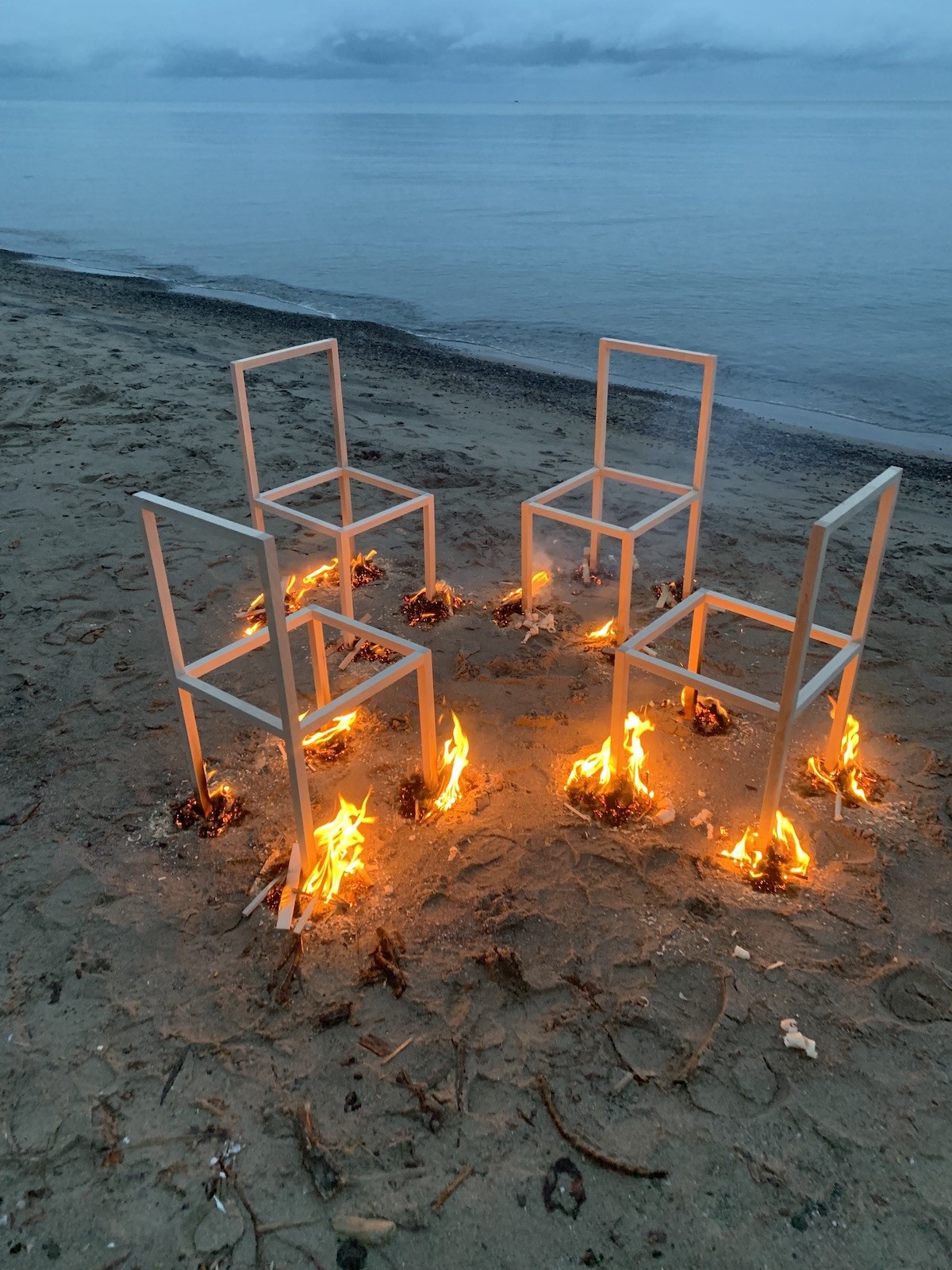 a group of five wood chairs that are on fire arranged in a circle on a sandy beach with blue water along the shore.