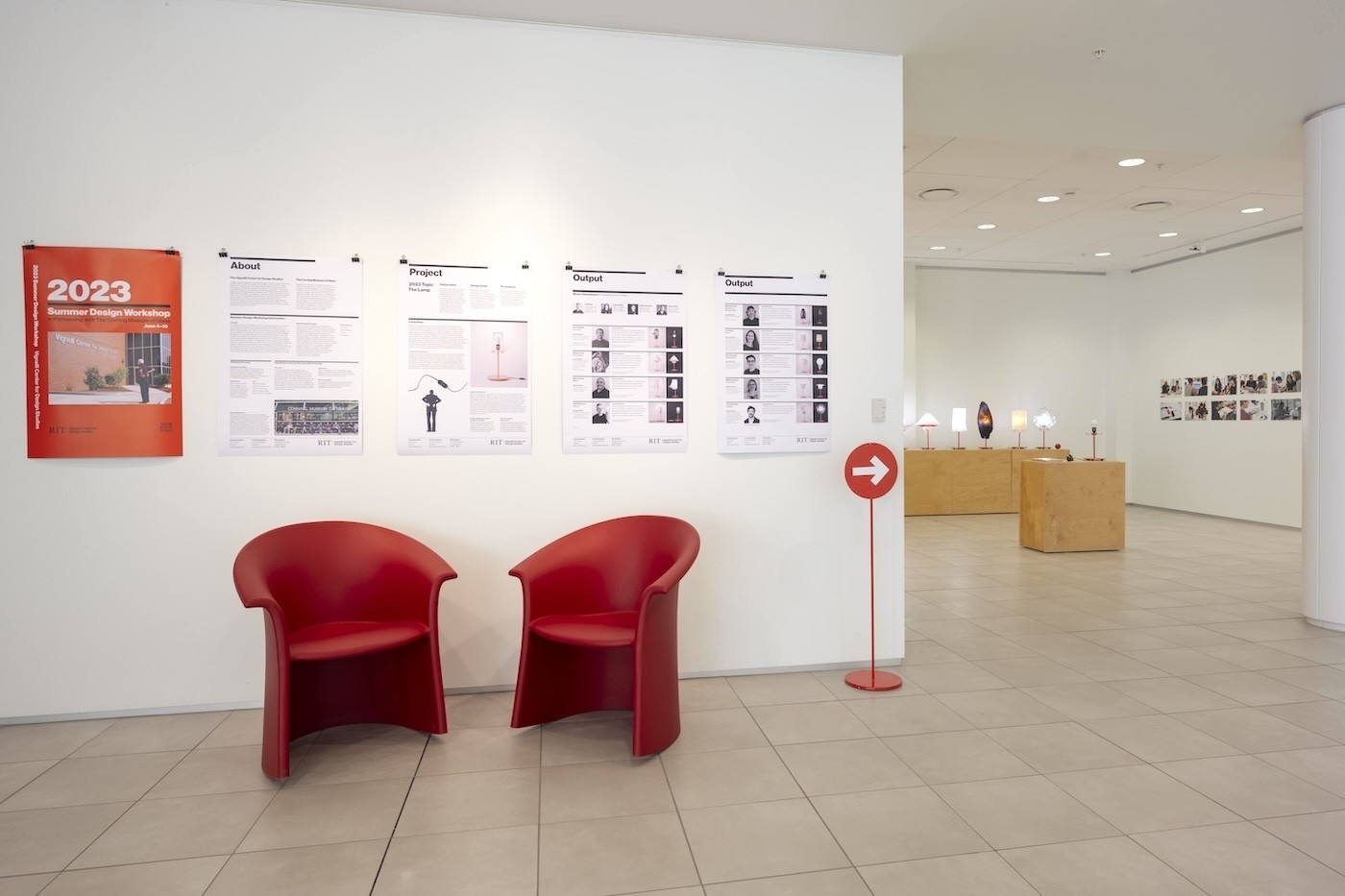 a view into the Grid Space gallery with two red chairs, four information panels and a view of the display of lamps on wood stands.