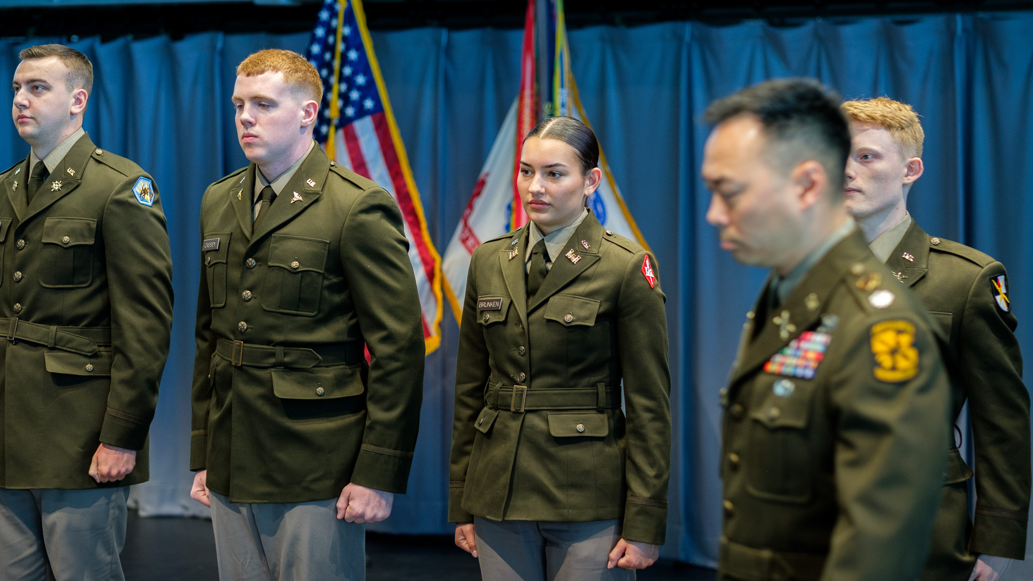 A group of cadets in dress uniform, standing at attention, with the US flag and Army flag behind them